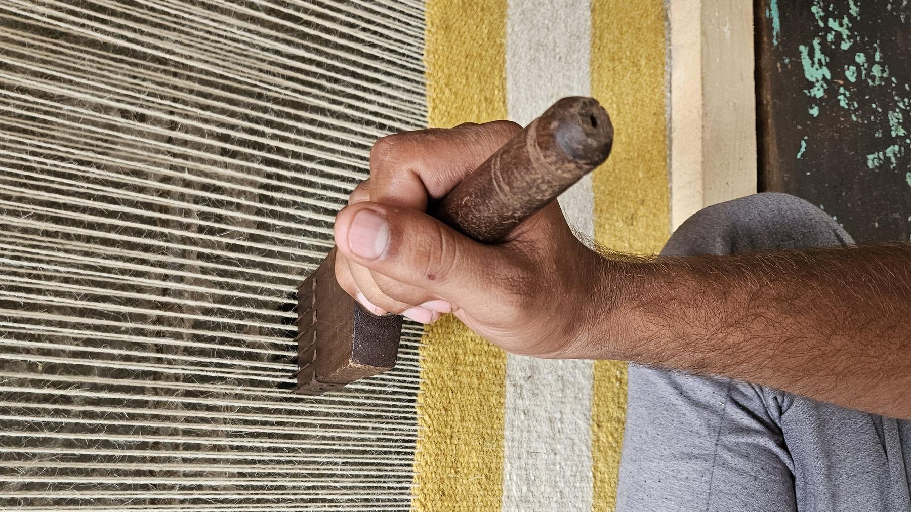 A weaver's hand pressing a beater comb into the warp of a yellow-striped flatweave rug on the loom in the Rug Gossips factory, Lahore